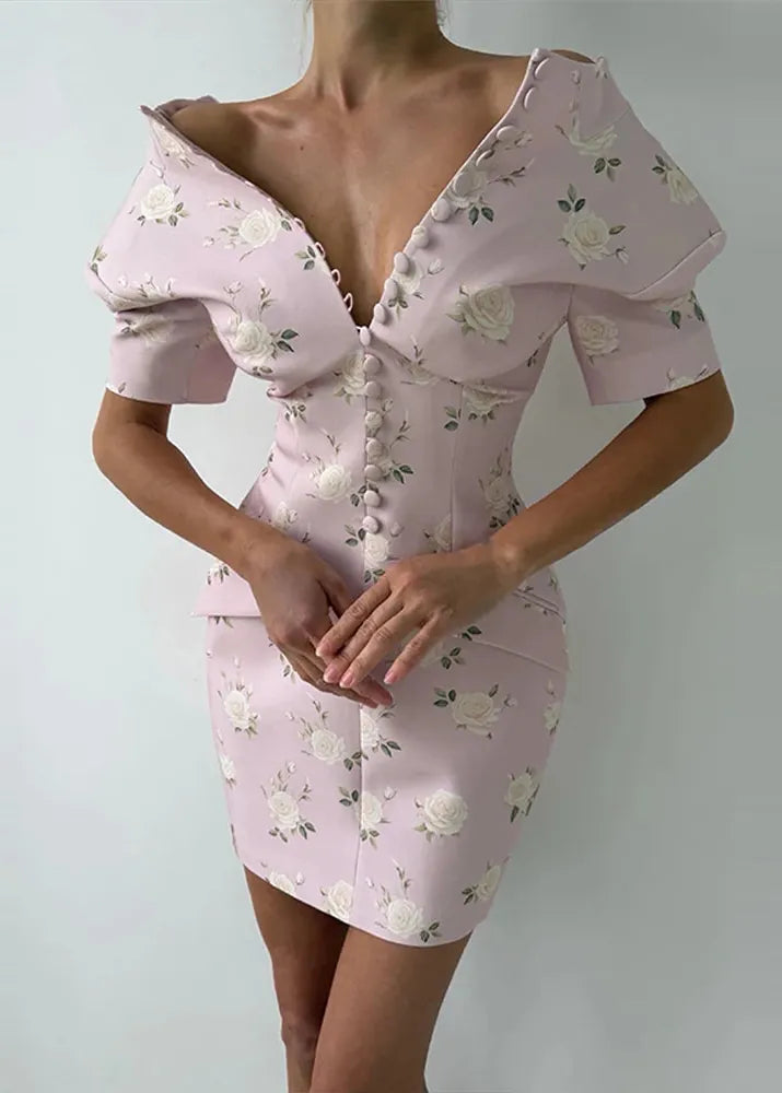 A woman wearing a light pink off-the-shoulder mini dress with a white rose floral pattern, featuring a deep plunging V-neckline and decorative buttons down the front.