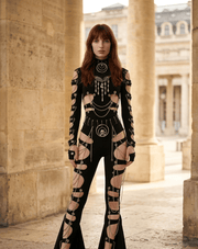 A model stands in a stone colonnade wearing a black long-sleeve flared jumpsuit heavily embellished with silver chains, rings, and cutout details that expose the skin. The Arvin logo is visible in the bottom right corner.
