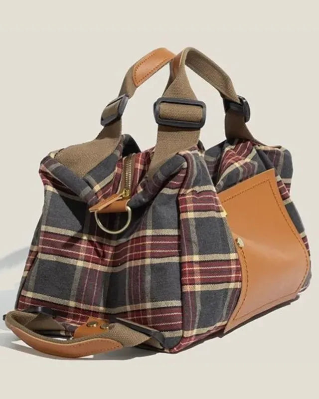 A duffel bag with a plaid pattern in navy, red, and beige, accented with tan leather panels and handles, shown on a light beige background.