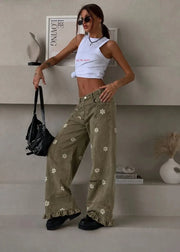 A woman models a pair of wide-leg olive green pants decorated with white floral prints and ruffled hems, paired with a white crop top and black sunglasses, standing on a minimalist concrete step platform.