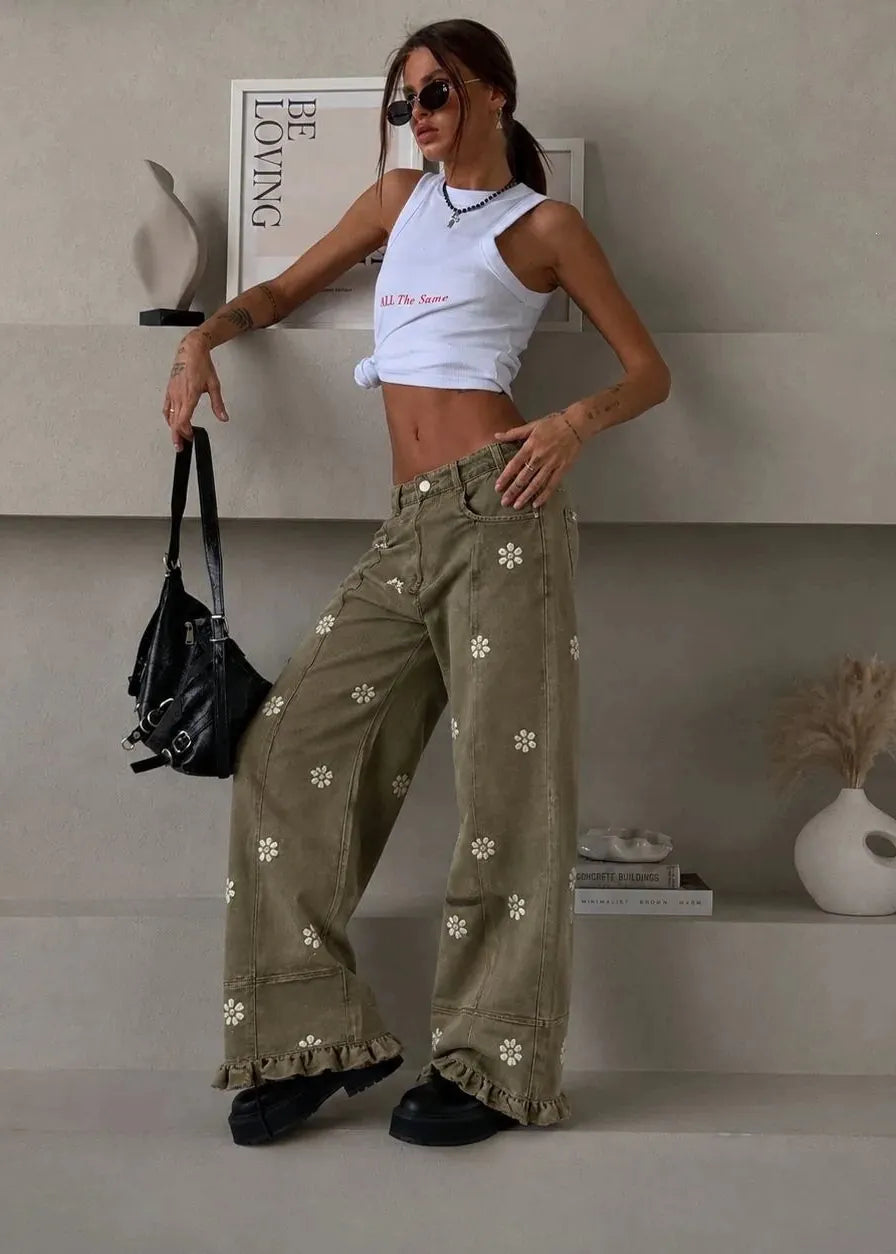 A woman models a pair of wide-leg olive green pants decorated with white floral prints and ruffled hems, paired with a white crop top and black sunglasses, standing on a minimalist concrete step platform.