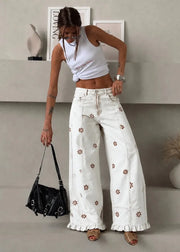 A woman models a pair of white wide-leg denim pants decorated with small brown floral embroidery and ruffled hems, styled with a white crop top and holding a black handbag.