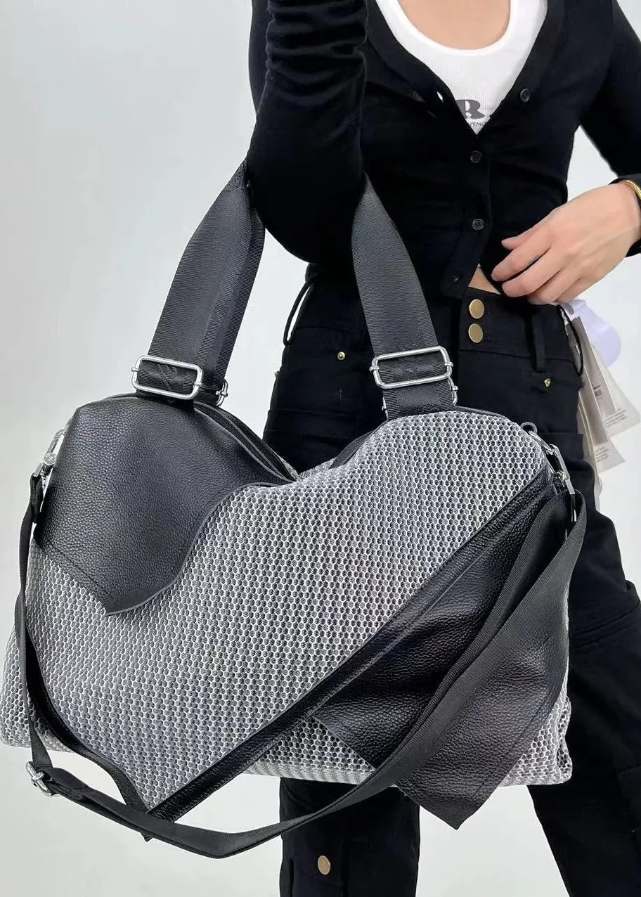A person wearing a black zip-up hoodie and black pants carries a large black and gray patterned duffel bag with a shoulder strap against a plain light gray background.