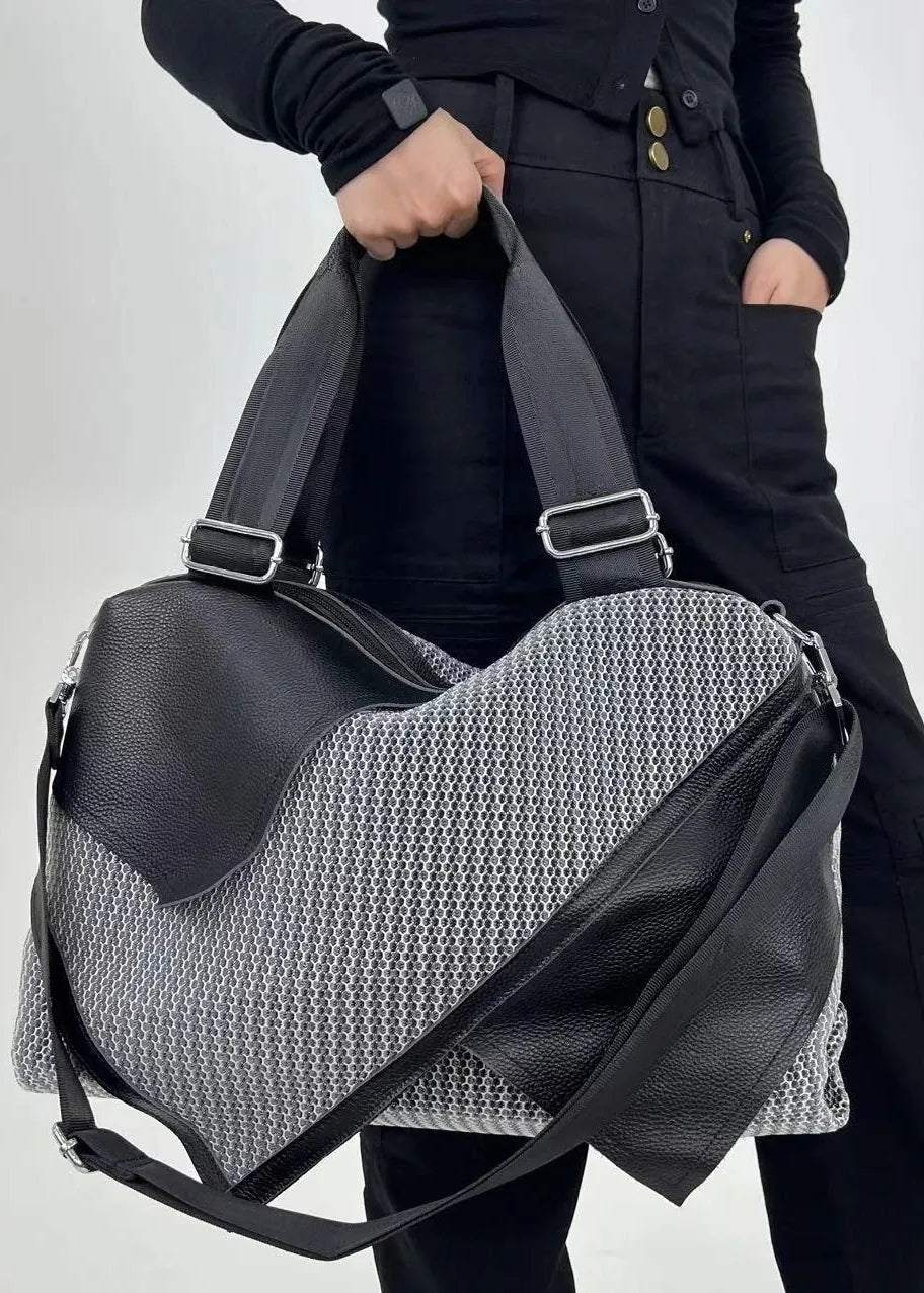 A person in black clothing holds a large black and gray patterned duffel bag with black leather-like panels and a black shoulder strap against a light gray studio background.
