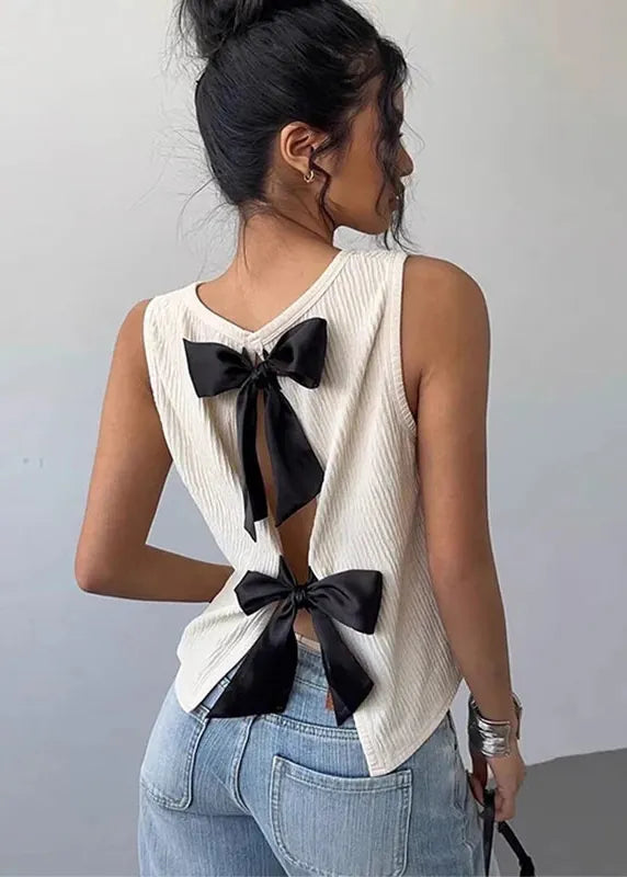 A woman shown from the back wearing a sleeveless cream ribbed top with large black bow details at the back, paired with light blue jeans, against a plain light gray wall.