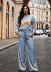 A woman stands on a city street under a stone archway wearing a light blue satin-like jumpsuit with gold embroidery and trim. The background shows a European-style street scene with buildings and parked cars.