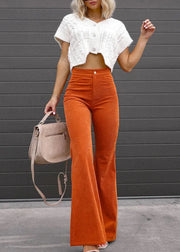 Woman wearing a white cropped top and orange flared pants, holding a beige handbag against a gray wall.