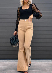Woman wearing a black top and beige pants with sheer sleeves against a gray wall.