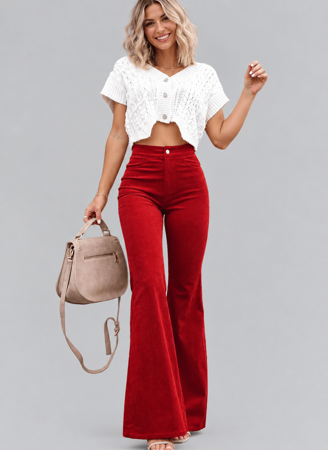 A woman modeling a white short-sleeve cropped knit top and red high-waisted flare pants, holding a taupe handbag against a solid gray studio background.
