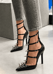 A person wearing black pointed-toe high heels with multiple silver metal spikes and cutout straps, paired with gray jeans in an indoor showroom setting.