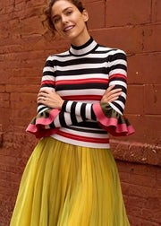 A woman wearing a black, white, and red striped mock-neck top with pink ruffle details on the sleeves, paired with a pleated yellow skirt, posing against a red brick wall.