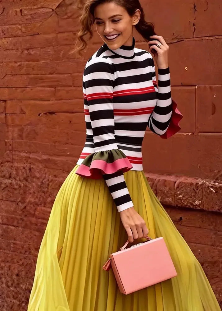 A woman posing in front of a rustic red brick wall, wearing a black, white, and red striped mock-neck top with ruffled details, a voluminous yellow pleated skirt, and holding a pink handbag.