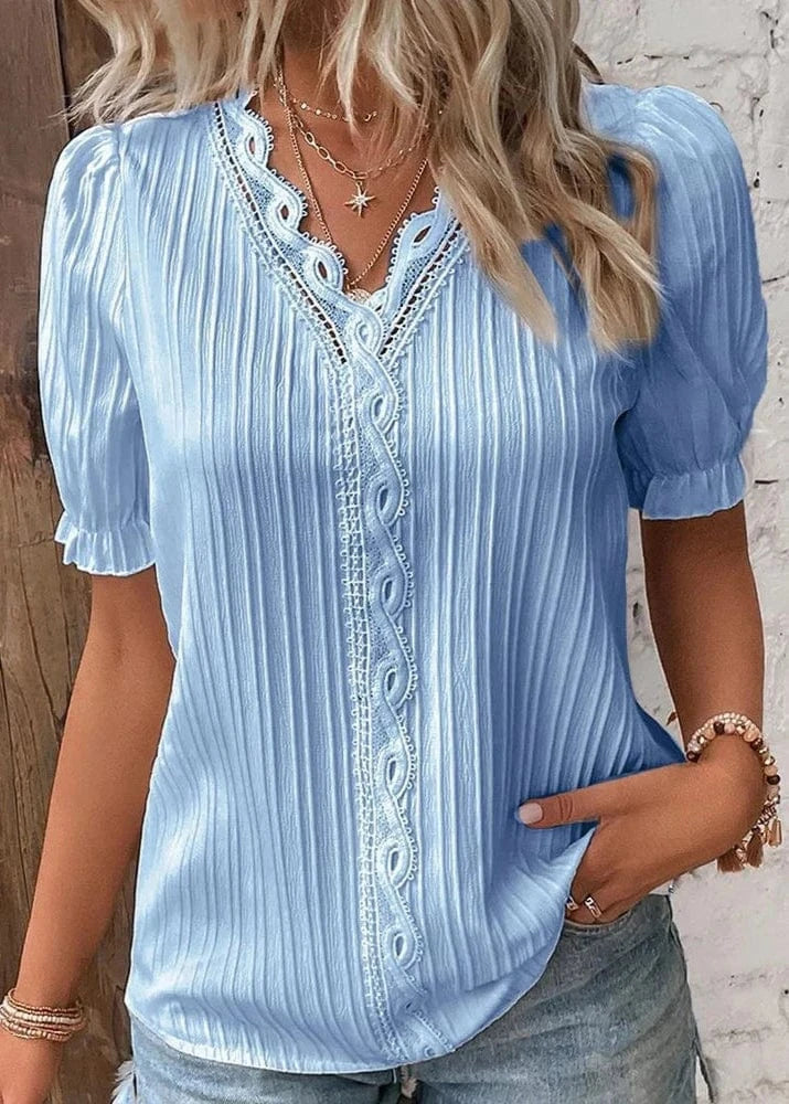 Woman wearing a light blue blouse with lace details against a wooden and stone wall.