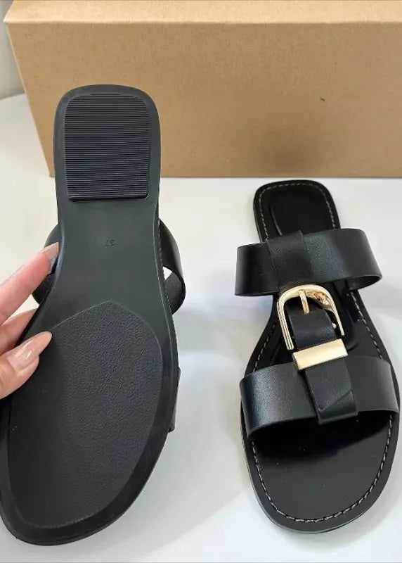 Black sandal with gold buckle held by a hand, next to a beige box on a white surface.