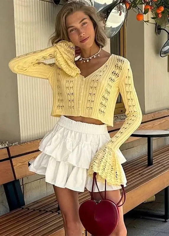 Woman wearing a yellow lace top and white skirt, holding a red heart-shaped bag, standing on a wooden bench.