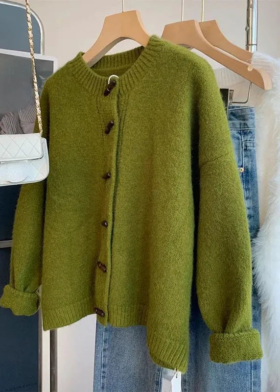 An olive green knit cardigan sweater with dark brown buttons, hanging on a wooden hanger on a clothing rack next to blue jeans, a white quilted shoulder bag, and a white faux fur garment.