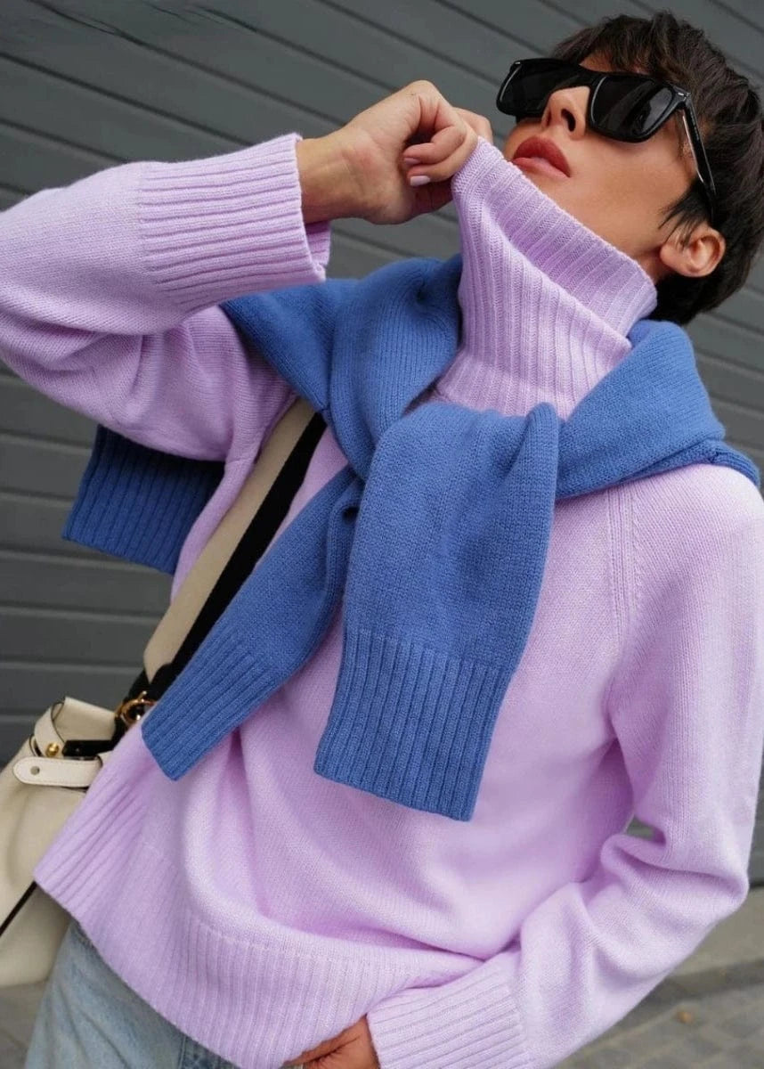 Person wearing a light purple sweater and blue cardigan with sunglasses, standing against a gray background.