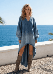 A woman stands outdoors by the ocean wearing a loose, long-sleeve high-low denim dress in faded blue with brown slouchy boots. The background shows a white wall, blue sea, and clear sky.