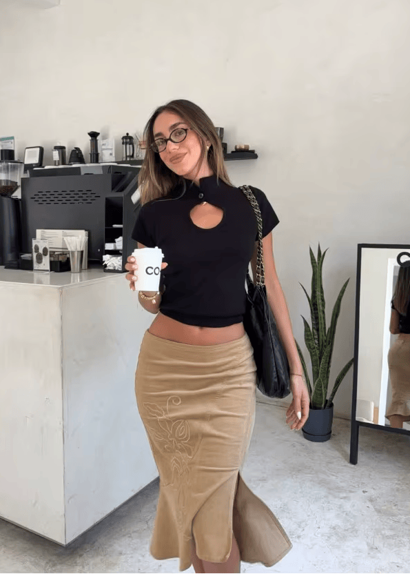 Woman in a black top and beige skirt holding a coffee cup in a modern cafe.