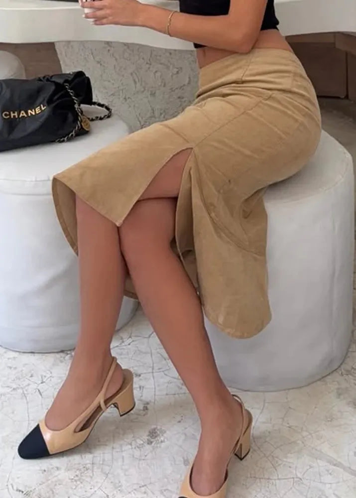 Person wearing a beige skirt and shoes sitting on a white stool with a Chanel bag in the background.