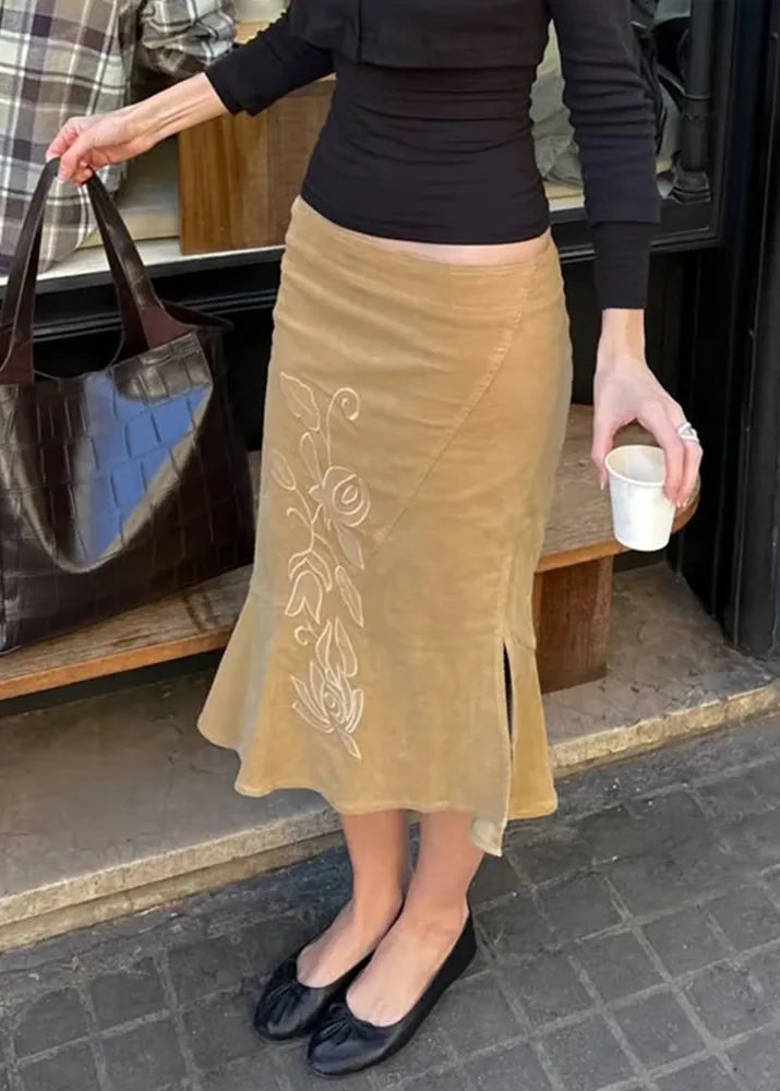 Person wearing a beige skirt with floral embroidery, holding a cup and a bag, standing on a sidewalk.