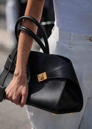 A close-up of a person carrying a structured black leather handbag with a prominent gold-tone turn-lock clasp and double top handles. The person is wearing a white top and white trousers, with the handbag featured prominently against a softly blurred outdoor background.