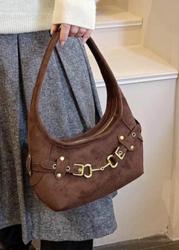 A person wearing a cream knit sweater and a gray wool skirt carries a brown suede shoulder bag with gold-tone hardware and horsebit details, standing indoors near white stairs.