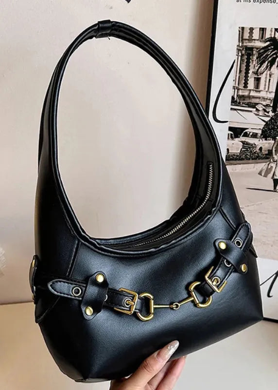 A black faux-leather hobo shoulder bag with gold-tone hardware, held by a hand against a neutral indoor background with a lamp and framed print.