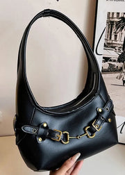 A black faux-leather hobo shoulder bag with gold-tone hardware, held by a hand against a neutral indoor background with a lamp and framed print.