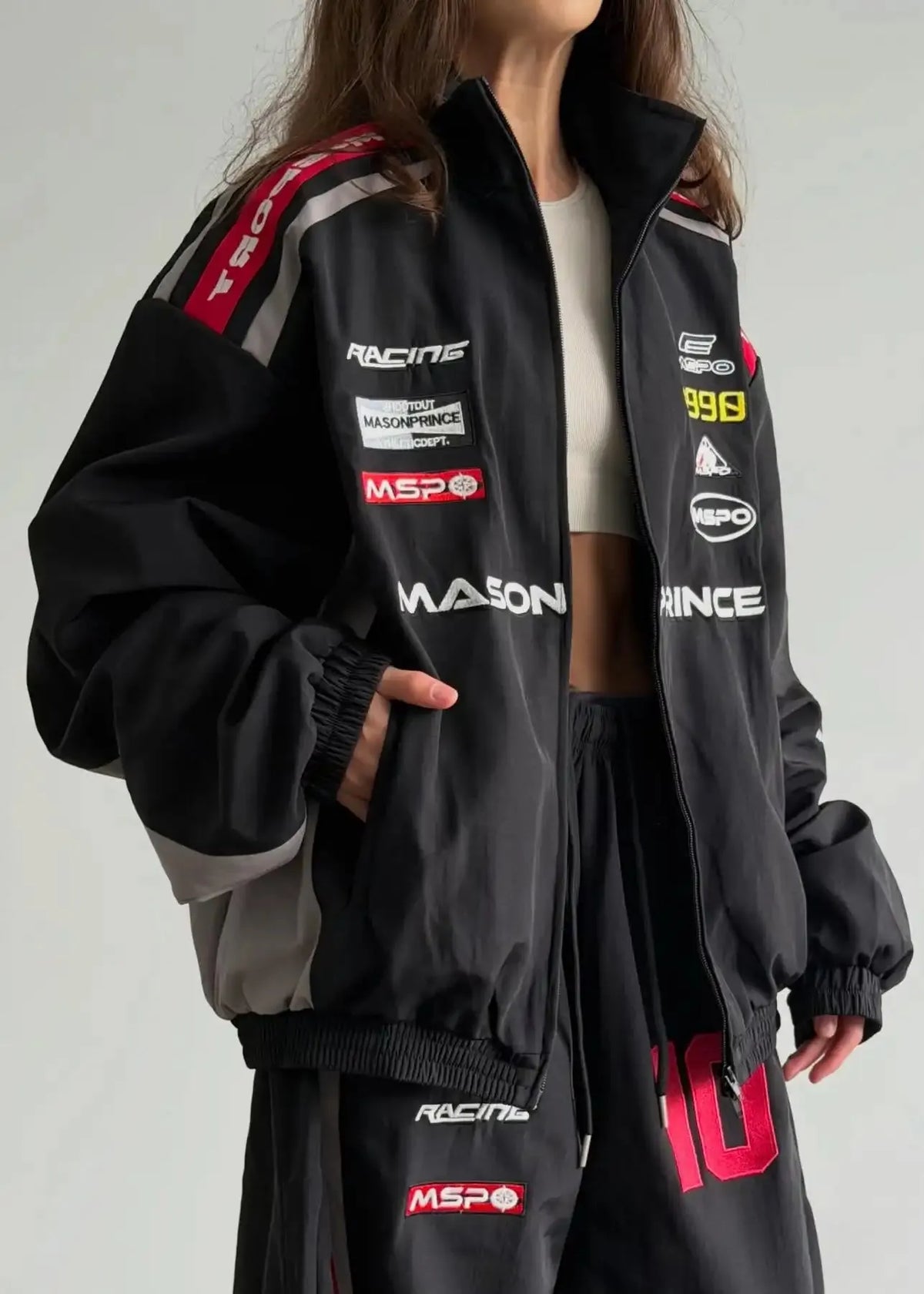 A person wearing an oversized black racing-inspired jacket with multiple sponsor-style logos and graphics in red, white, and gray, shown against a plain light background.