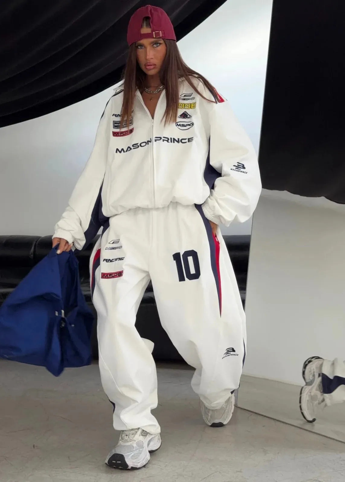A woman models a white racing-inspired tracksuit with 'MASON PRINCE' and sponsor logos, paired with a burgundy cap and holding a blue bag.