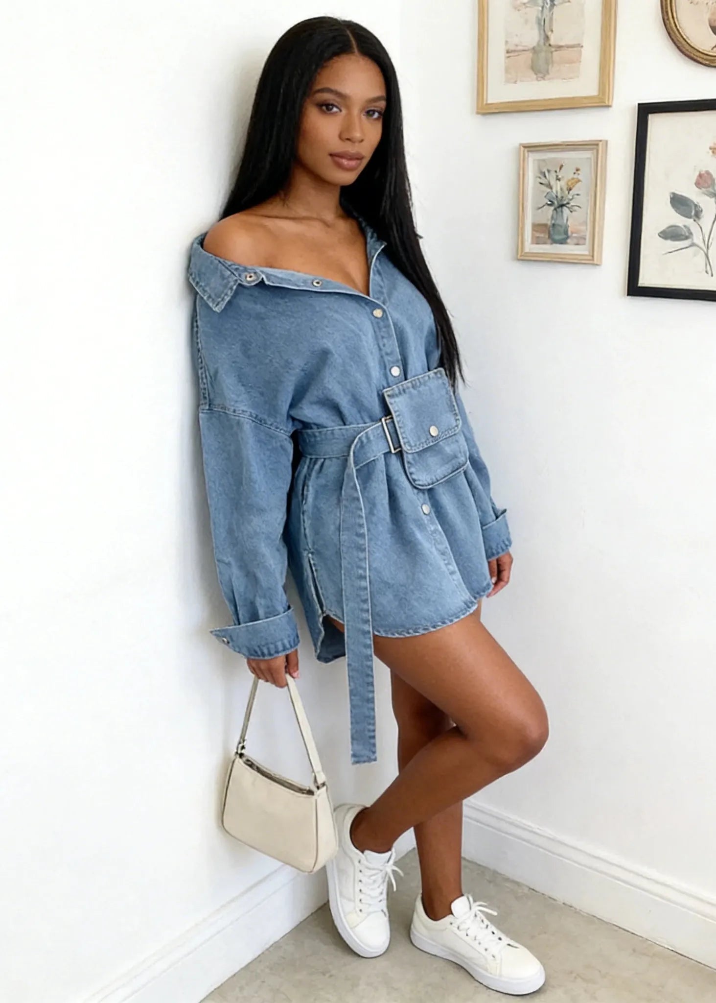Woman wearing a denim dress with white sneakers in a room with framed pictures on the wall.