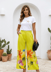 Woman wearing a white t-shirt and yellow floral pants standing in front of a white archway with potted plants.