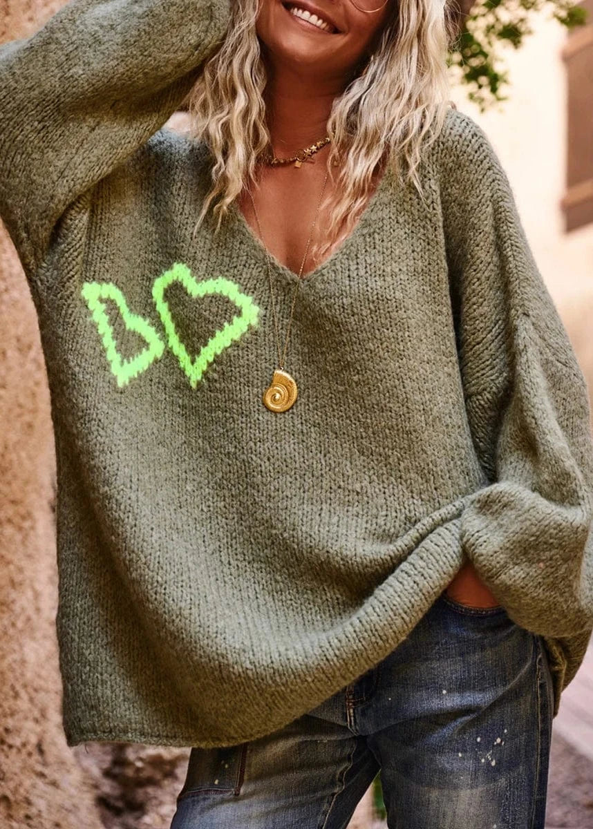Person wearing a green sweater with heart designs and a button, standing against a blurred background.